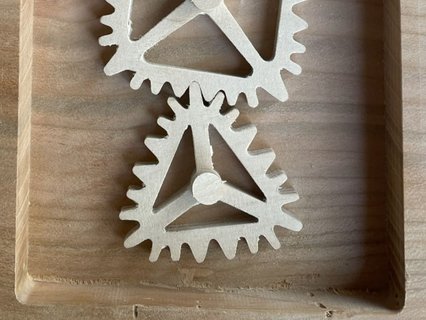 Holzmechanik mit drei ineinandergreifenden, unregelmäßigen Zahnrädern in runder, quadratischer und dreieckiger Form. Die Zahnräder sind in einem rechteckigen Holzrahmen montiert und lassen sich manuell drehen. Das Objekt demonstriert spielerisch die Funktionsweise nicht‑kreisförmiger Getriebe.