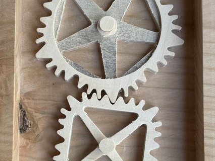 Holzmechanik mit drei ineinandergreifenden, unregelmäßigen Zahnrädern in runder, quadratischer und dreieckiger Form. Die Zahnräder sind in einem rechteckigen Holzrahmen montiert und lassen sich manuell drehen. Das Objekt demonstriert spielerisch die Funktionsweise nicht‑kreisförmiger Getriebe.